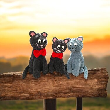 Drei gehäkelte Amigurumi-Katzen (zwei schwarz mit roten Schleifen, eine graublaue) sitzen auf einer Holzlatte vor Sonnenuntergang