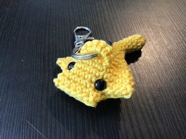 Gehäkelter gelber Amigurumi-Schlüsselanhänger mit schwarzen Augen und Ohren auf dunklem Tisch