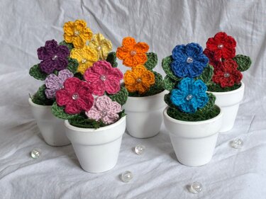 Close-up of several small white pots each holding a colorful crochet flower with green leaves and a clear bead, arranged on a white fabric background.