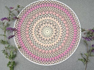 Rundes gehäkeltes Mandala mit Farbverlauf von Beige zu Rosa auf weißem Ring, dekoriert mit lila Wildblumen auf grauem Hintergrund.