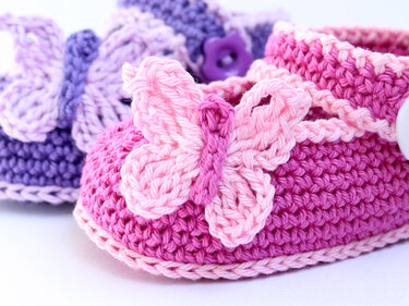 Nahaufnahme eines gehäkelten rosa Babyschuhs mit Schmetterlings-Applikation, violetter Babyschuh im Hintergrund