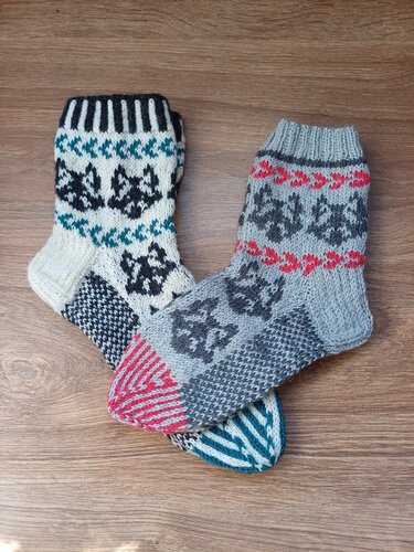 Zwei gestrickte Socken mit Tiermotiven in Grau, Weiß, Rot und Türkis auf Holzhintergrund.