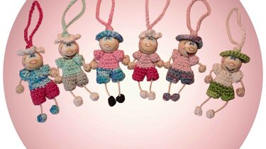 Sechs gehäkelte Figuren mit Holzperlenköpfen, bunten Mützen und Kleidern, die sich an den Händen halten vor rosafarbenem Hintergrund.