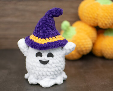 Weißes gehäkeltes Amigurumi-Geistchen mit violettem Hexenhut und gelbem Zierstreifen, Kürbisse im Hintergrund