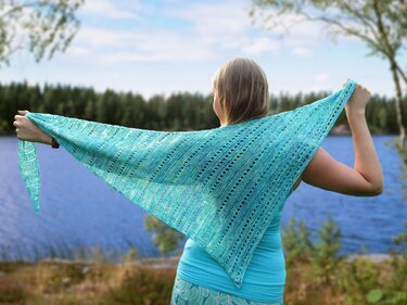 Person hält türkisfarbenes gestricktes Dreieckstuch mit Lochmuster vor einem See