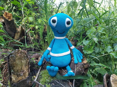 Blaues gehäkeltes Alien-Amigurumi mit großen Augen sitzt auf einem Baumstumpf im Grünen.