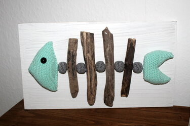 Gehäkeltes Fischskelett aus Treibholz und grauen Häkelkugeln auf weißem Holzbrett.