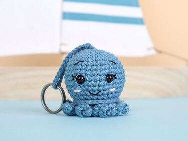 Blue crocheted octopus amigurumi keychain with a smiling embroidered face, black safety eyes, and a metal ring on a light blue surface.