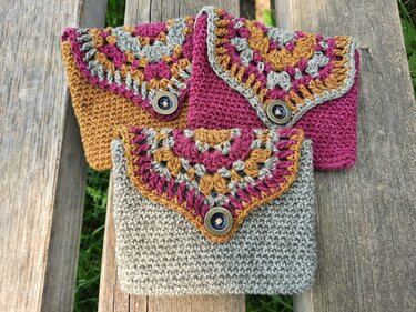 Drei gehäkelte Täschchen in Senf, Pink und Hellgrau mit mehrfarbigen Granny‑Klappen und Knopfverschlüssen auf einer Holzbank