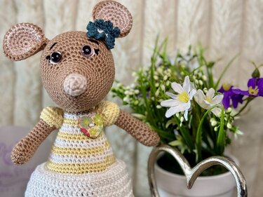 Gehäkelte beige Amigurumi-Maus mit gelb-weiß gestreiftem Kleid, Herzknopf und Häkelblume neben Topf mit weißen und violetten Blumen
