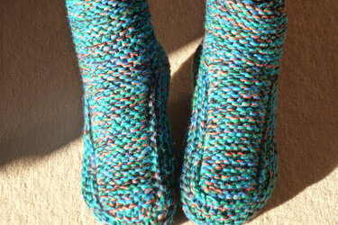 Nahaufnahme eines Paars gestrickter mehrfarbiger Hausschuhe (türkis, grün, orange) auf beigem Teppich im Sonnenlicht.