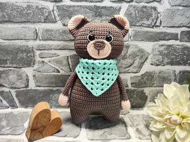 Brauner Amigurumi-Teddybär mit mintgrünem Halstuch und beigen Details vor grauer Ziegelwand