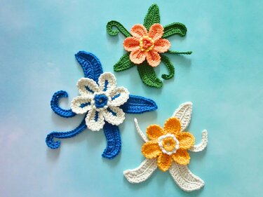 Drei gehäkelte Blumen-Applikationen in Orange, Blau und Gelb mit Blättern auf hellblauem Hintergrund.