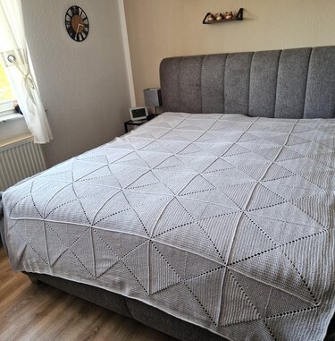 Hellgraue gehäkelte Tagesdecke mit Dreiecksmuster auf einem Bett mit grauem Kopfteil.