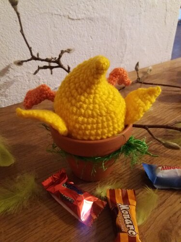 Gehäkelter gelber Eierwärmer in Kükenform im Tontopf mit Ostergras und Weidenkätzchen