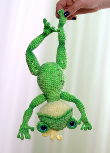 Gehäkelter grüner Amigurumi-Frosch hängt kopfüber an den Beinen, große blaue Augen