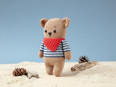 Light brown crocheted teddy bear wearing a blue-and-white striped shirt and red bandana standing on sand with pinecones and driftwood.