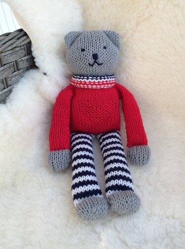 Gestrickter grauer Teddybär mit rotem Herzpullover und schwarz-weiß gestreiften Beinen auf hellem Fell