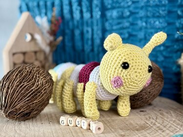 Gehäkelte Amigurumi-Raupe mit gelbem Kopf und bunten Segmenten auf Holzoberfläche