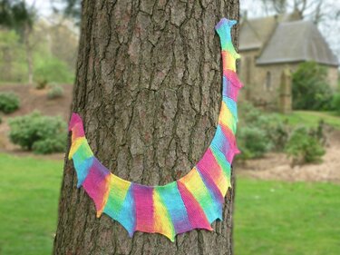Knitted rainbow dragon tail scarf wrapped around a tree trunk, with a blurred building and lawn in the background.
