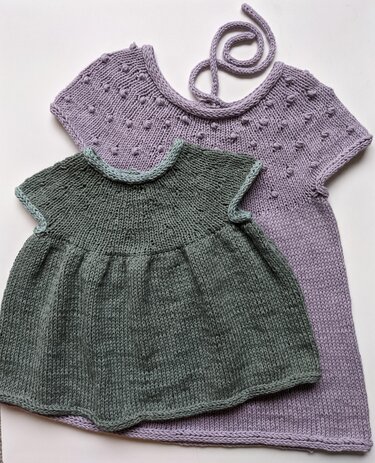 Two knitted children's dresses: a small green A-line baby dress with gathered skirt in front and a longer lavender dress with bobble-stitch yoke.