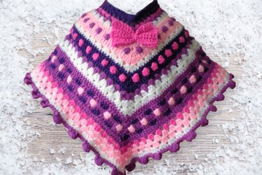 Gehäkelter Poncho für Kinder in Lila, Rosa und Weiß mit Zierschleife auf hellem Hintergrund.