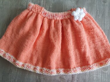 Pfirsichfarbener gestrickter Babyrock mit weißer Häkelblüte und pastellfarbener Abschlusskante