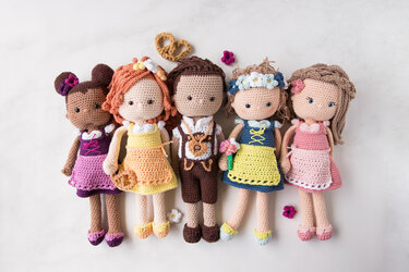 Fünf gehäkelte Amigurumi‑Puppen nebeneinander in bunten Kleidern, eine mit lederhosenartiger Tracht