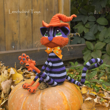 Purple and black striped crocheted cat amigurumi with an orange witch hat and bow, seated on a pumpkin.