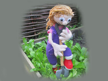 Amigurumi gardener doll in purple overalls and red boots holding a shovel in a raised garden bed.