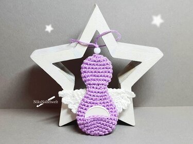 Lila gehäkelter Engel mit weißen Flügeln an weißem Holzstern vor grauem Hintergrund