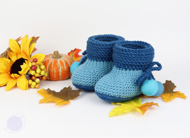 Soft blue crochet baby booties with felt pom-poms beside a pumpkin, sunflower, and fall leaves