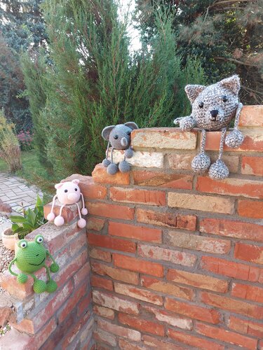 Vier gehäkelte Amigurumi auf Ziegelmauer: grüner Frosch, rosa Schwein, grauer Koala, graue Katze