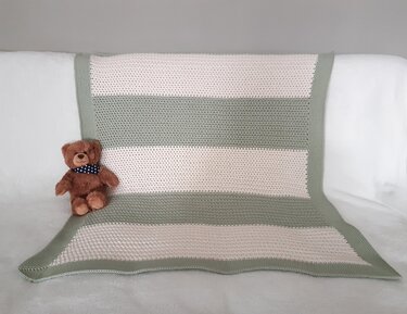 Gehäkelte Babydecke mit breiten hellgrünen und cremefarbenen Streifen, dekorativ drapiert neben einem braunen Teddybär