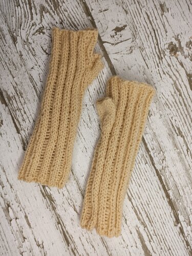 Zwei gestrickte fingerlose Handschuhe in Beige mit Rippenmuster auf hell gestrichener Holzfläche.
