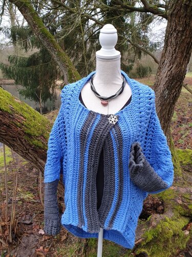 Gehäkelte Damenjacke blau-grau mit Streifen und Schneeflockenbrosche auf Schneiderpuppe vor Wald