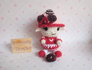 Red-and-white amigurumi lamb wearing a dress and matching hat with a small crocheted ball in front.
