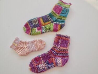Drei gestrickte Kindersocken: eine rosa Babysocke mit Knöpfen und zwei bunte Mustersocken auf weißem Grund.