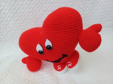Rotes gehäkeltes Amigurumi-Herz mit aufgenähten Augen, lächelndem Mund, kleinen Armen und weißen Sohlen auf hellem Hintergrund.