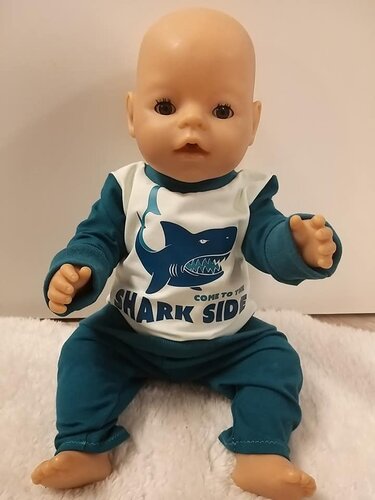 Sitzende Babypuppe trägt weißes Shirt mit blauem Hai-Aufdruck und dem Schriftzug 'Shark Side', dazu petrolfarbene Hose und Ärmel.