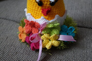 Gehäkeltes gelbes Amigurumi-Küken mit rotem Schnabel, weißer Eierschale, buntem Blumenkranz und rosa Schleife, Nahaufnahme