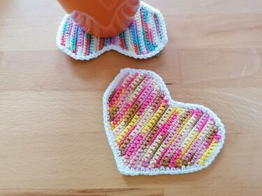 Zwei bunte herzförmige gehäkelte Untersetzer auf Holz, einer unter einer orangefarbenen Tasse