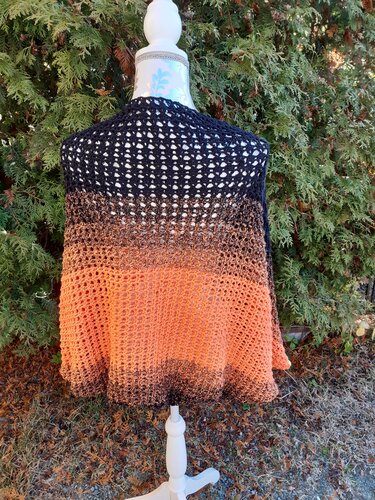 Rückenansicht eines gehäkelten Seelenwärmers in Schwarz‑Braun‑Orange auf einer Schneiderpuppe vor grünen Zweigen