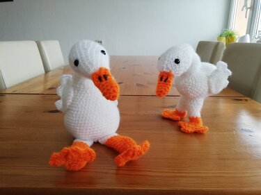 Zwei weiße gehäkelte Enten-Amigurumi mit orange Schnäbeln und Füßen auf Holztisch