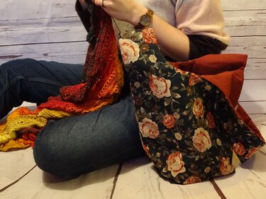Buntes Häkelstück neben gemusterter Stofftasche mit Rosenprint und rotem Innenfutter auf Holzfußboden