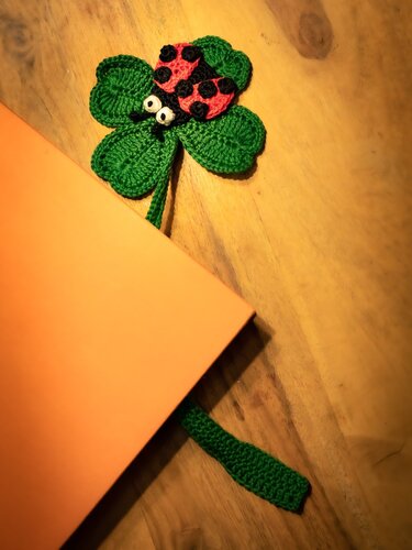 Grünes gehäkeltes vierblättriges Kleeblatt mit rotem Marienkäfer, als Lesezeichen in einem orangefarbenen Buch.