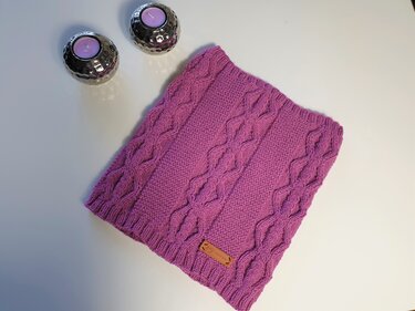 Magenta Loop‑Schal mit Zopfmuster und kleinem Lederetikett, auf weißer Fläche neben zwei silbernen Kerzenhaltern mit lila Kerzen.