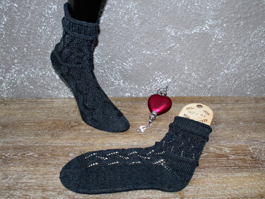 dunkelgraue gestrickte Socken mit Ajourmuster; eine auf Sockenspanner (Gr. 40/41), eine auf Fußpuppe; roter Herzanhänger