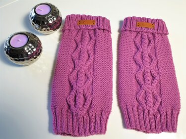 Zwei lila gestrickte Beinstulpen mit Zopfmuster auf weißem Untergrund neben zwei silbernen Kerzenhaltern mit lila Teelichtern.