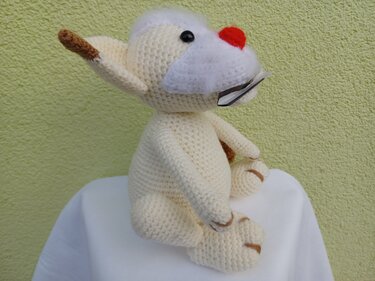 Beiges gehäkeltes Amigurumi-Haustier mit roter Nase, seitlich sitzend vor hellgrüner Wand